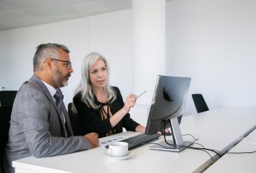 Focused business colleagues watching content on computer, pointing at display and talking while sitting at desk with paper chart. Business communication concept